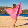 young woman walking bareboot on golden sand towards the blue calm sea holding a dark pink striped hammam towel