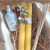 set of three items on a craft box. A reed diffuser with the sticks and a set of two beeswax candle sticks tied with thread and a beautiful bunch of white dried flowers